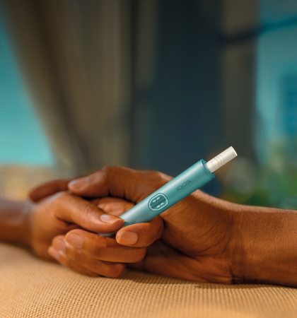 A woman holds an IQOS device while looking at a laptop.